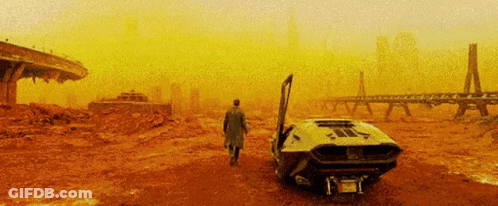 A Man In A Trench Coat Is Walking Towards A Car With Its Door Open In The Desert . GIF
