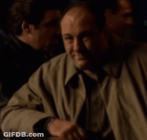 A Man In A Trench Coat Sits At A Table With Other Men GIF
