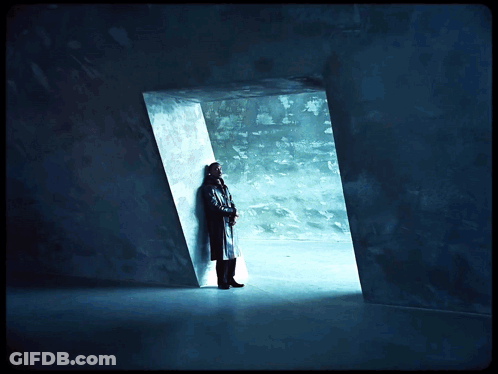 A Man In A Trench Coat Stands In A Dark Room Looking Out A Doorway GIF
