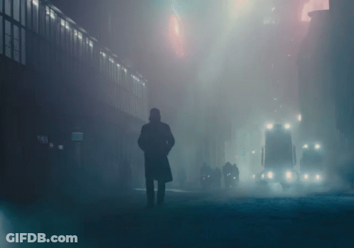 A Man In A Trench Coat Stands In A Foggy City Street GIF