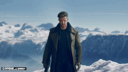 A Man In A Trench Coat Stands On Top Of A Snow Covered Mountain With The Name Pranay Varma Below Him GIF