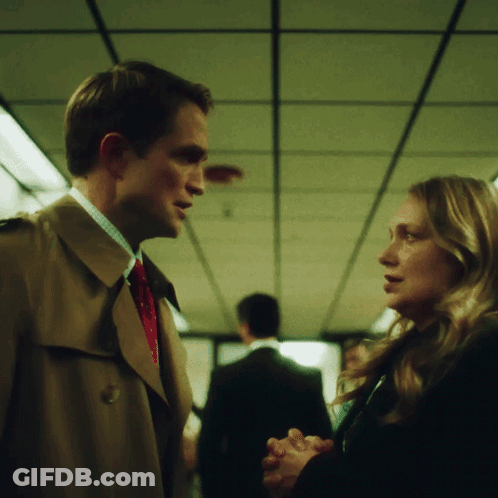 A Man In A Trench Coat Talks To A Woman In A Black Coat GIF
