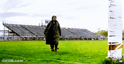 A Man In A Trench Coat Walks Across A Field With A Stadium In The Background GIF