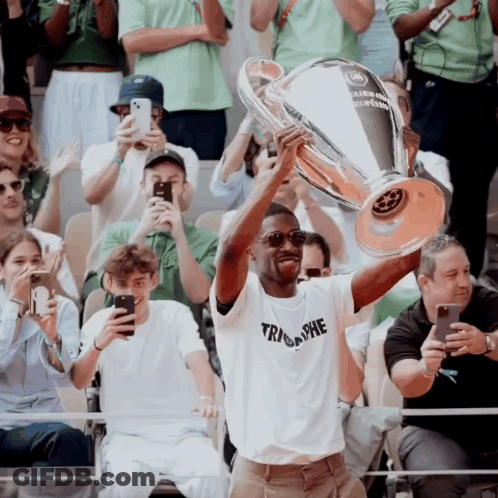 A Man In A Triumphe Shirt Holds Up A Trophy In Front Of A Crowd Of People GIF