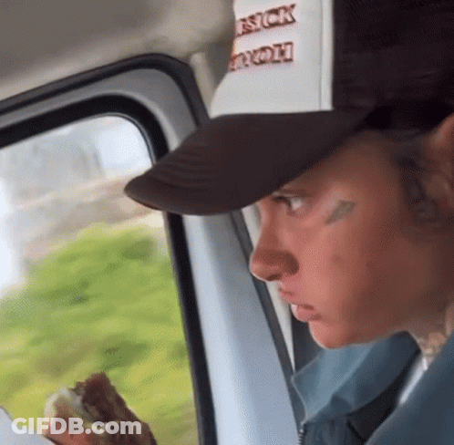 A Man In A Trucker Hat Is Eating A Hamburger While Looking Out The Window . GIF