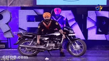 A Man In A Turban Is Standing On A Motorcycle In Front Of A Sign That Says " Real " GIF