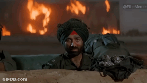 A Man In A Turban Looks Out From Behind A Wall With A Fire In The Background GIF