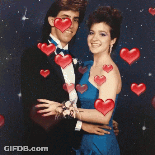 A Man In A Tuxedo And A Woman In A Blue Dress Are Posing For A Picture With Red Hearts Surrounding Them GIF