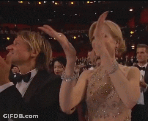 A Man In A Tuxedo And A Woman In A Dress Are Clapping Their Hands In A Crowd . GIF