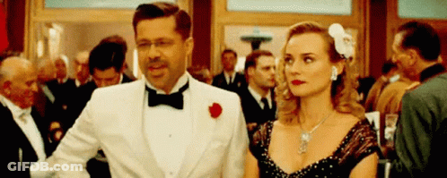 A Man In A Tuxedo And A Woman In A Dress Are Walking In A Crowd . GIF