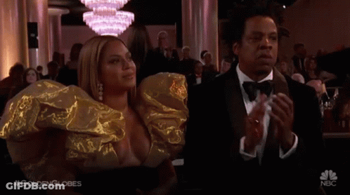 A Man In A Tuxedo And A Woman In A Gold Dress Are Applauding At A Golden Globes Event . GIF