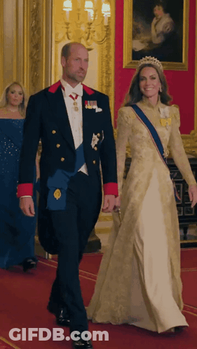 A Man In A Tuxedo And A Woman In A Gown Are Walking Down A Red Carpet GIF
