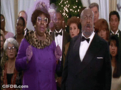 A Man In A Tuxedo And A Woman In A Purple Coat Stand In Front Of A Crowd Of People GIF