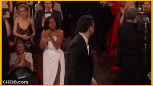 A Man In A Tuxedo And A Woman In A White Dress Are Standing Next To Each Other In Front Of A Crowd . GIF