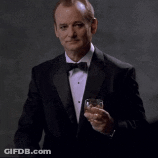 A Man In A Tuxedo And Bow Tie Is Holding A Glass Of Whiskey And Pointing At The Camera . GIF