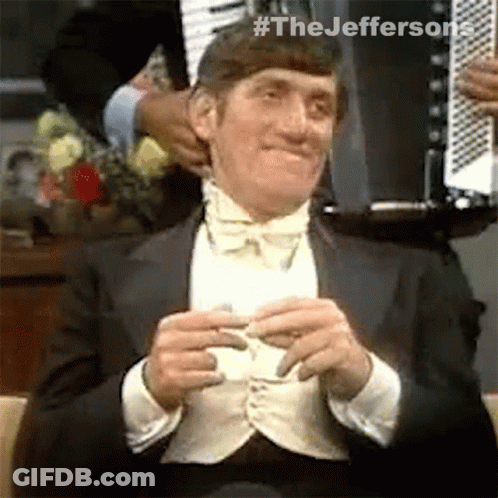 A Man In A Tuxedo And Bow Tie Is Sitting In A Chair And Holding A Piece Of Paper . GIF