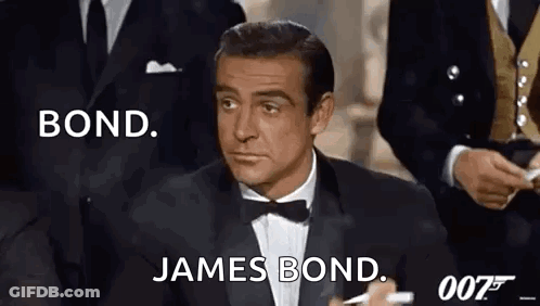 A Man In A Tuxedo And Bow Tie Is Sitting In Front Of A Group Of Men In Suits . GIF