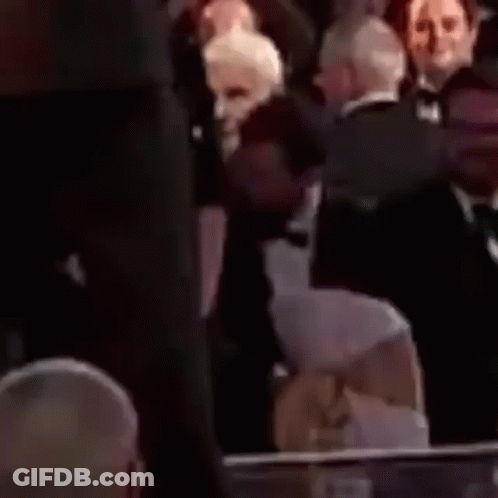 A Man In A Tuxedo And Bow Tie Is Sitting Next To A Woman In A Crowd . GIF