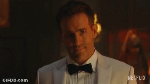 A Man In A Tuxedo And Bow Tie Is Smiling In A Dark Room . GIF