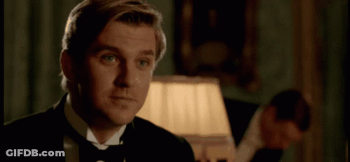 A Man In A Tuxedo And Bow Tie Is Smiling In A Room With A Lamp . GIF