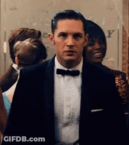 A Man In A Tuxedo And Bow Tie Is Standing In A Room With Other People . GIF