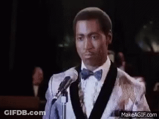 A Man In A Tuxedo And Bow Tie Is Standing In Front Of A Crowd Of People . GIF