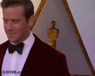 A Man In A Tuxedo And Bow Tie Is Standing In Front Of A Statue Of An Oscar Winner . GIF