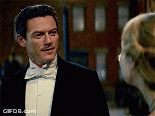 A Man In A Tuxedo And Bow Tie Looks At A Woman In A White Dress GIF
