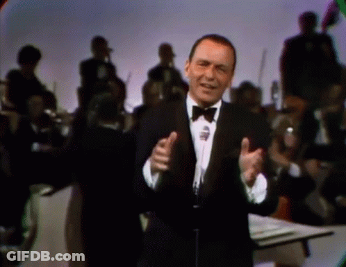 A Man In A Tuxedo And Bow Tie Singing Into A Microphone GIF