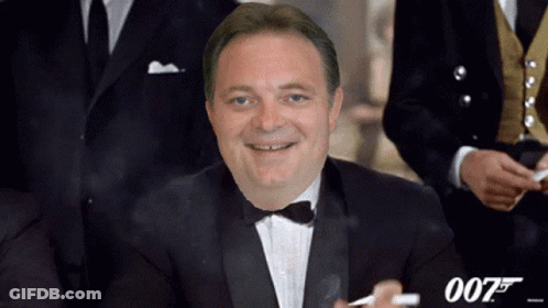 A Man In A Tuxedo And Bow Tie Smoking A Cigarette With The Number 007 Behind Him GIF