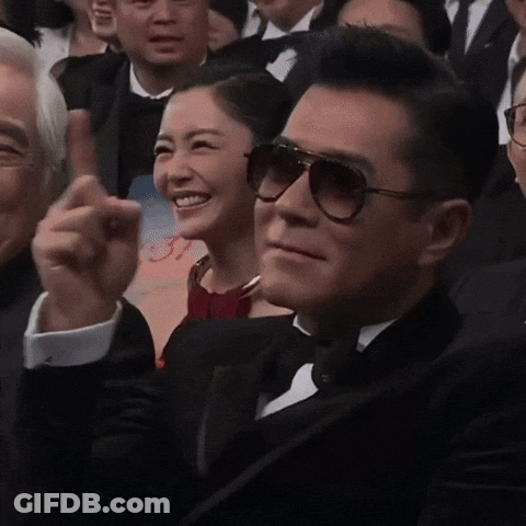A Man In A Tuxedo And Sunglasses Is Sitting In A Crowd GIF