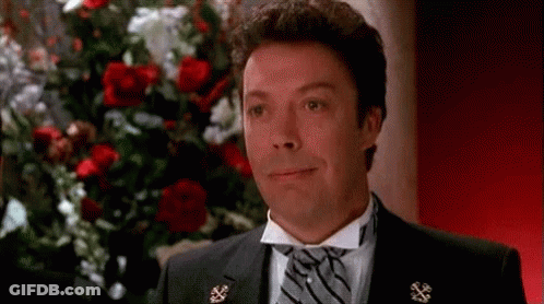 A Man In A Tuxedo And Tie Is Smiling In Front Of A Bunch Of Roses . GIF