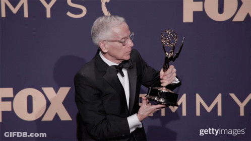 A Man In A Tuxedo Holds A Trophy In Front Of A Fox Advertisement GIF