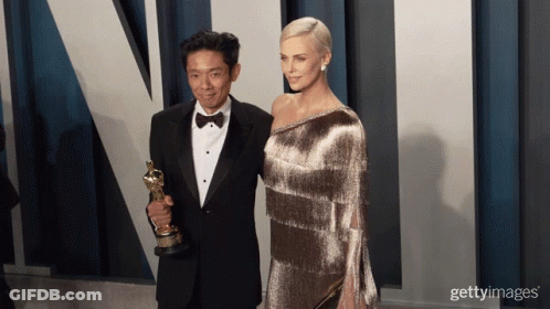 A Man In A Tuxedo Holds An Oscar Trophy Next To A Woman GIF