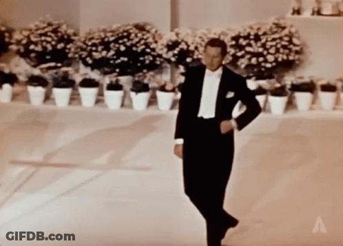 A Man In A Tuxedo Is Dancing In Front Of Potted Flowers GIF