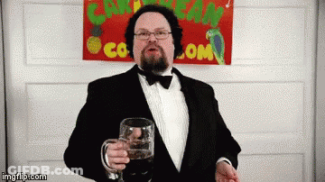 A Man In A Tuxedo Is Holding A Glass In Front Of A Sign That Says Caribbean Co.com GIF