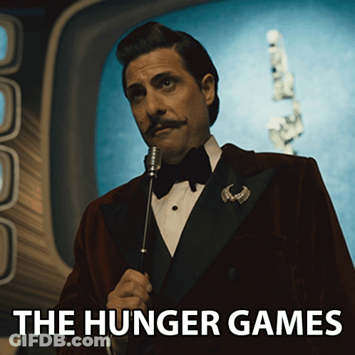 A Man In A Tuxedo Is Holding A Microphone And The Hunger Games Is Written Below Him GIF