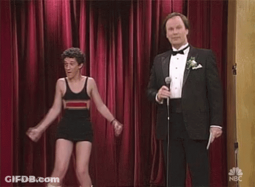 A Man In A Tuxedo Is Holding A Microphone While A Man In A Bathing Suit Is Dancing On Stage . GIF