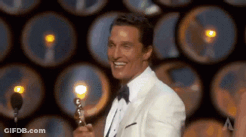 A Man In A Tuxedo Is Holding A Trophy And Smiling While Standing In Front Of A Microphone . GIF