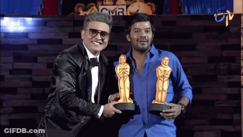 A Man In A Tuxedo Is Holding A Trophy Next To Another Man In A Blue Shirt GIF