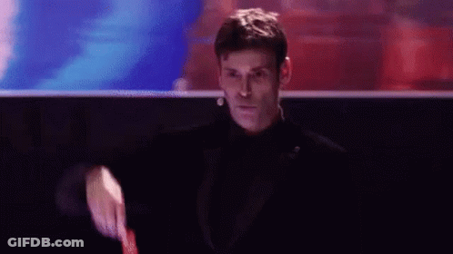 A Man In A Tuxedo Is Holding A Watch And Pointing At The Camera . GIF