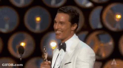 A Man In A Tuxedo Is Holding An Oscar Trophy On A Stage And Smiling . GIF