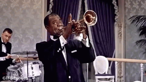 A Man In A Tuxedo Is Playing A Trumpet In A Room With A Drummer In The Background . GIF