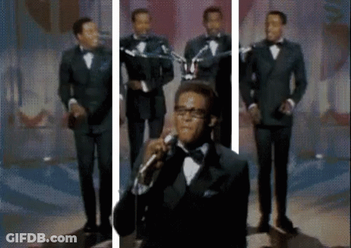 A Man In A Tuxedo Is Singing Into A Microphone In Front Of A Group Of Men . GIF