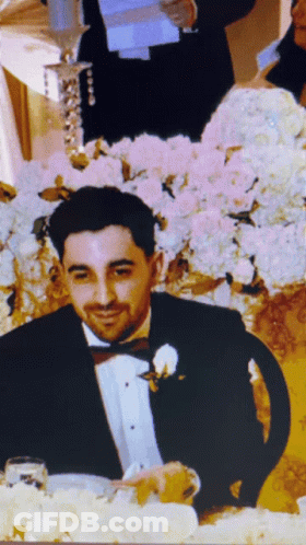 A Man In A Tuxedo Is Sitting At A Table With Flowers In The Background GIF