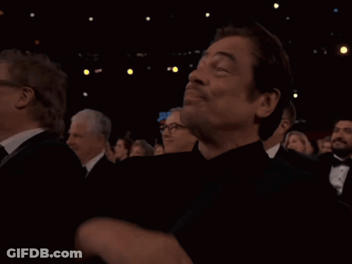 A Man In A Tuxedo Is Sitting In A Dark Room With Other People GIF
