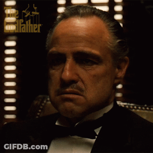 A Man In A Tuxedo Is Sitting In Front Of A Window With The Godfather Logo On It GIF