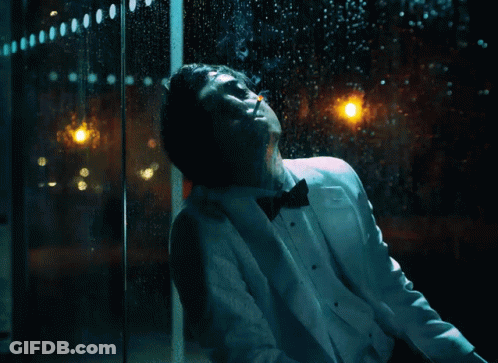A Man In A Tuxedo Is Smoking A Cigarette In Front Of A Window With Rain Drops On It GIF