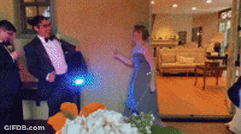 A Man In A Tuxedo Is Standing Next To A Woman In A Blue Dress In A Living Room GIF