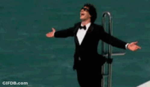 A Man In A Tuxedo Is Standing On A Boat With His Arms Outstretched . GIF